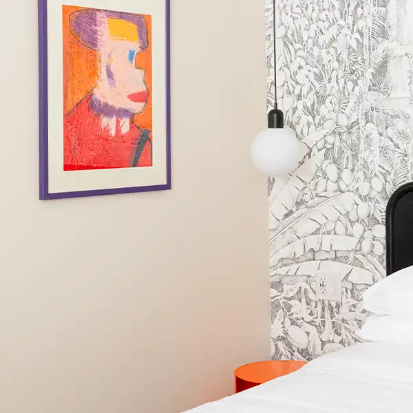 Beige room, with a bed with white sheets, a round hanging lamp, and a colorful painting