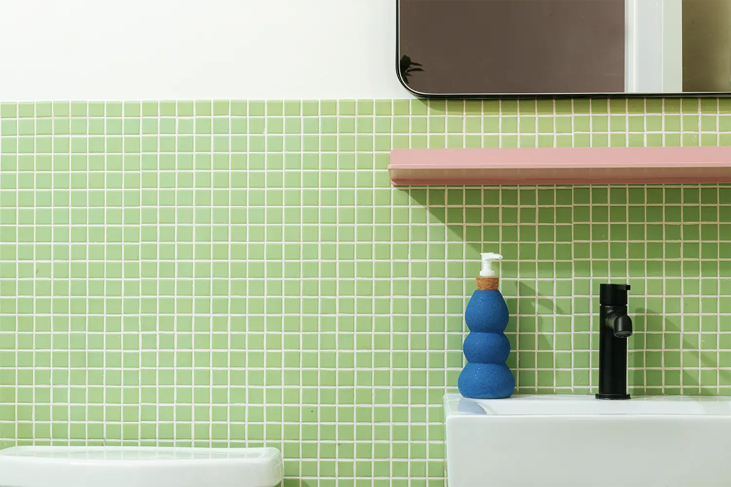 Bathroom corner with a blue hand soap dispenser on a white sink in front of a green tiled wall with a pink shelf above