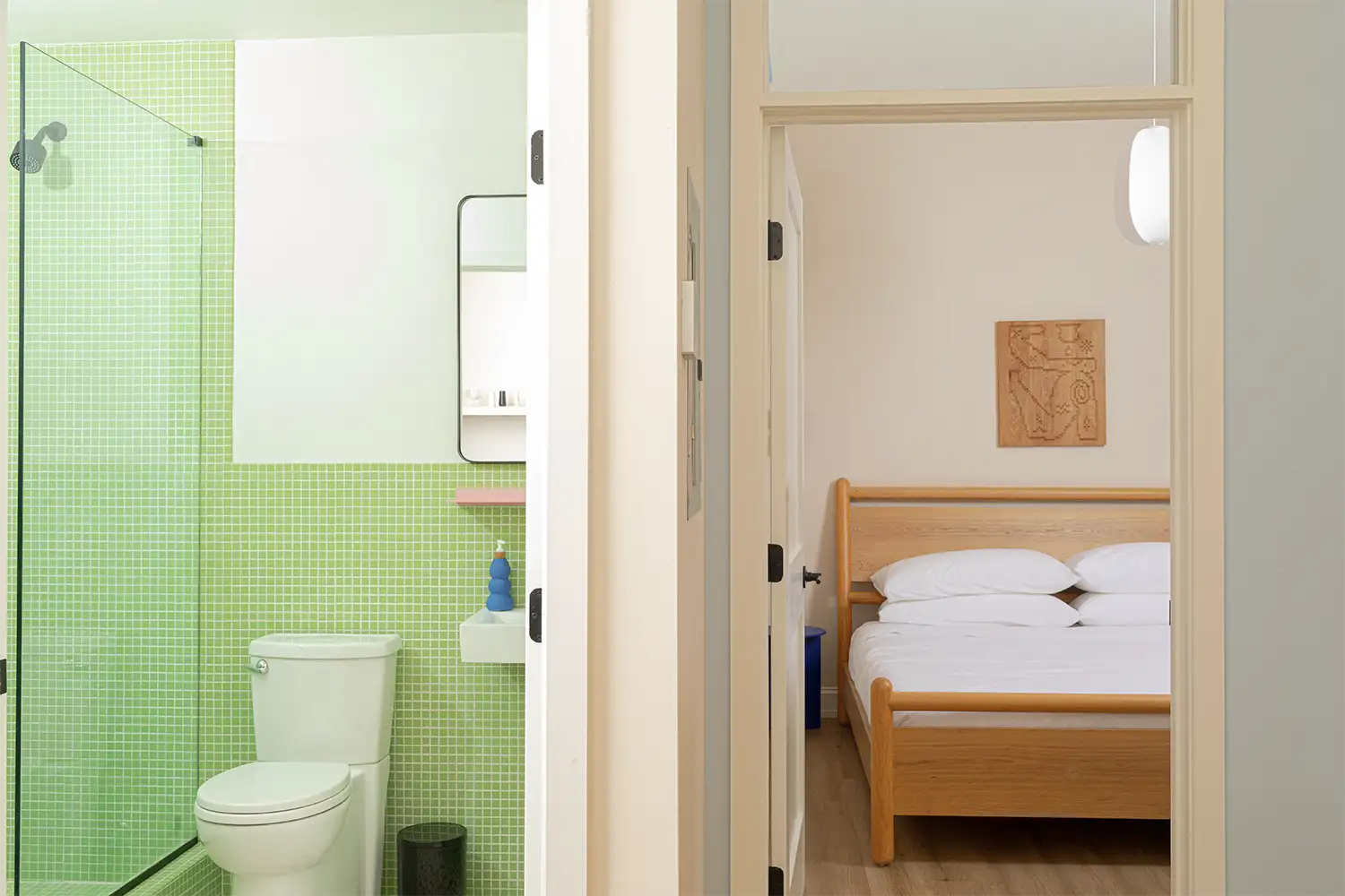 General view of the interior of a green bathroom with a white toilet and sink, separated by a wall from a bedroom with a double wooden bed and white bedding.