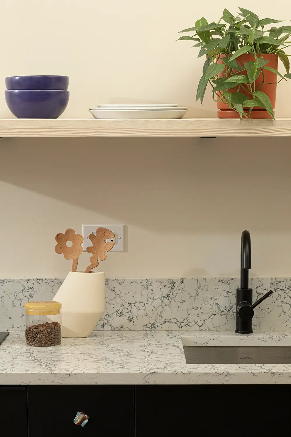 Close up of a marble kitchen counter with sink and kitchen utensils and a beige shelf on top with white plates, purple bowls and a plant