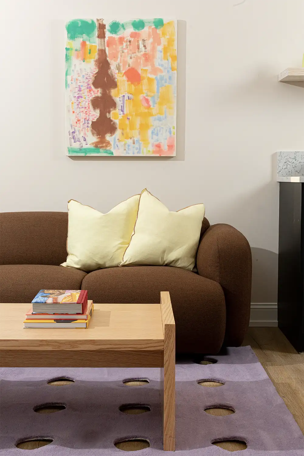 Beige living room whit a brown sofa with light yellow cushions, a wood coffee table, a purple modern carpet and a colorful painting hanging on the wall behind the sofa.