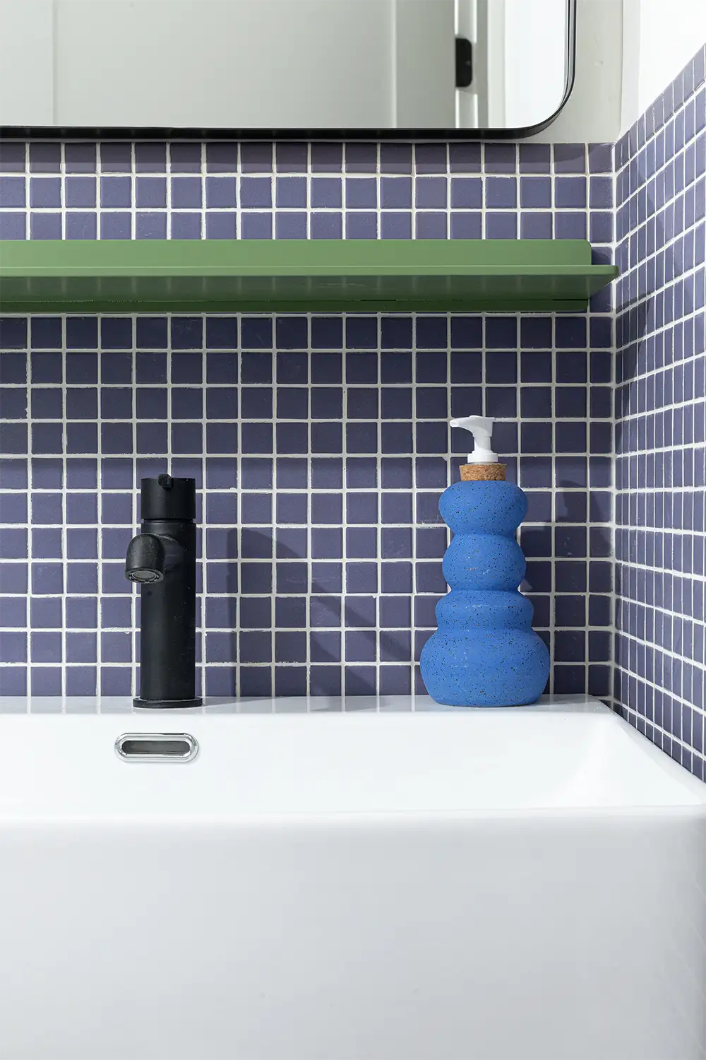 Bathroom corner with a blue hand soap dispenser on a white sink in front of a dark blue tiled wall with a green shelf above