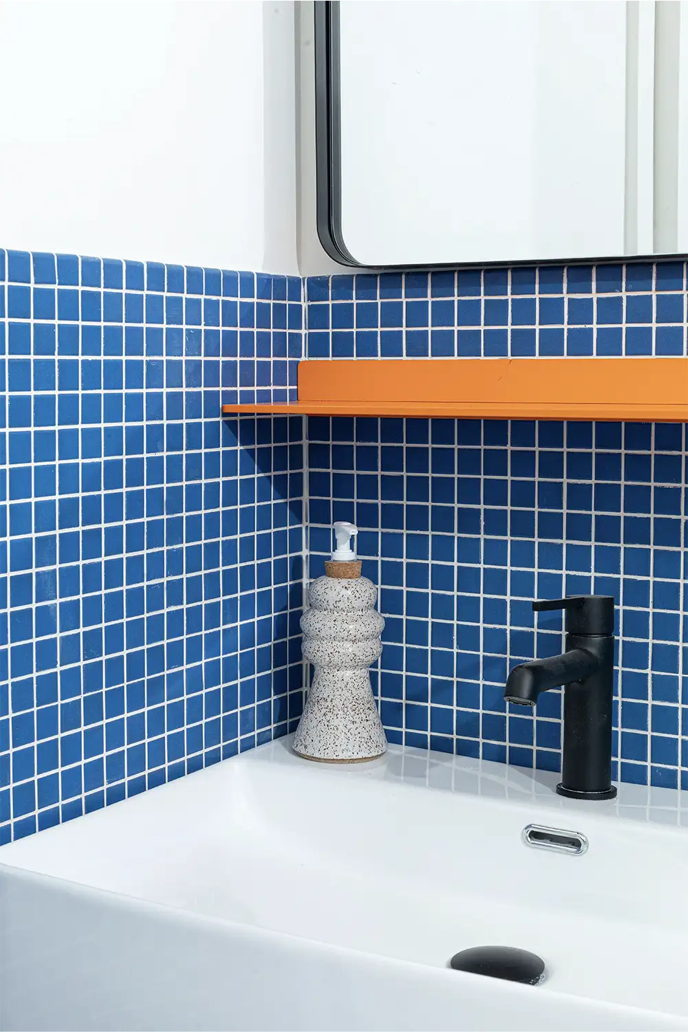 Bathroom with a white hand soap dispenser on a white sink in front of a royal-blue tiled wall and a mirror with an orange shelf beneath