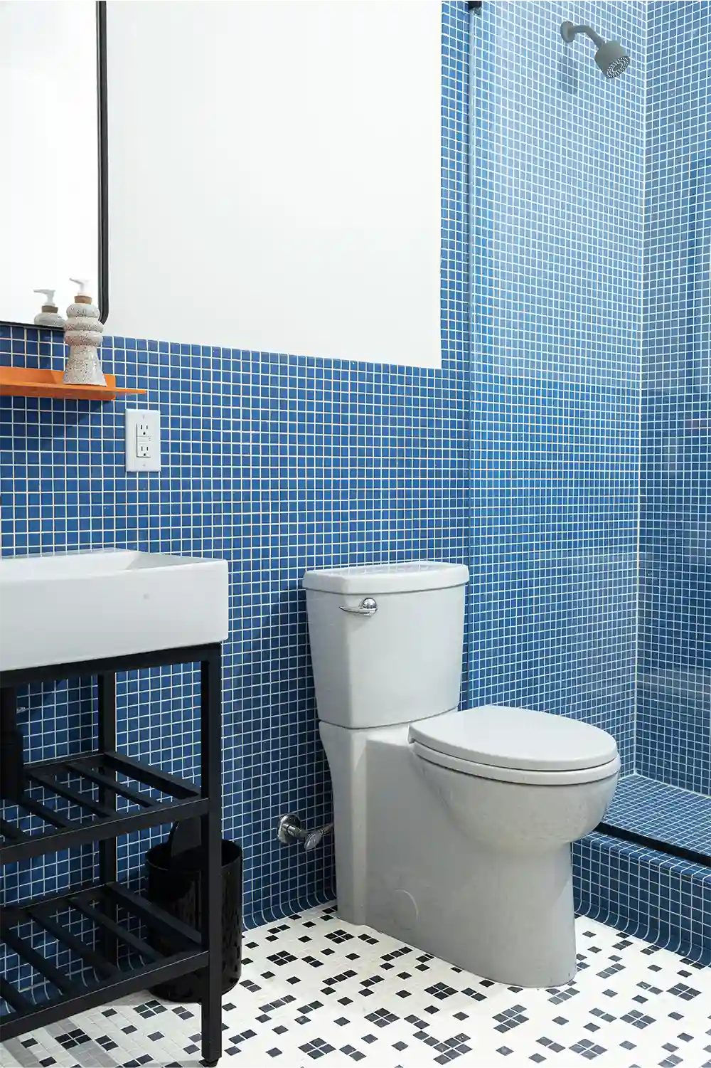 Bathroom with blue tiles, a large hanging mirror, a orange shelf underneath, a white sink with hand soap dispenser, a white toilet, and a glass-enclosed shower.