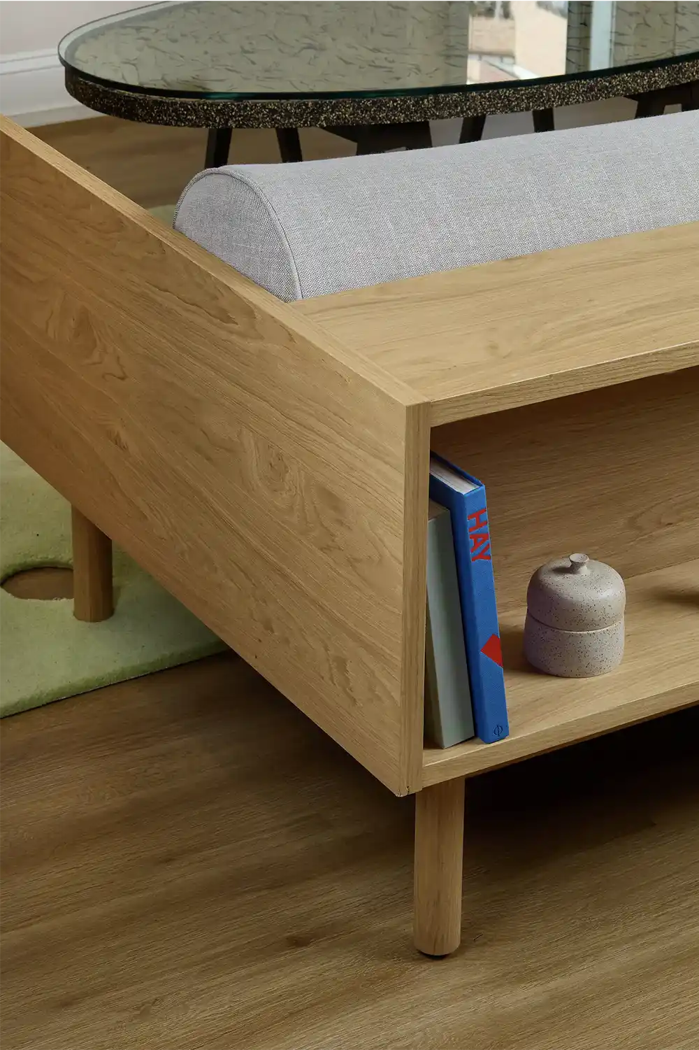 Close-up of a wooden sofa with built-in shelving holding books and a small ceramic object, placed on a wooden floor beside a glass coffee table.