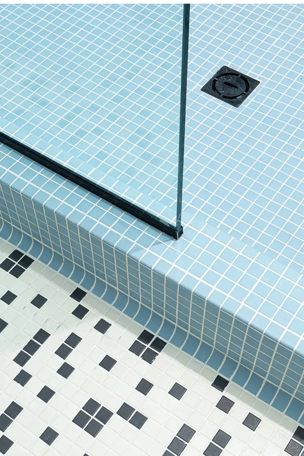 Modern bathroom design with pastel blue mosaic shower tiles and patterned black-and-white floor tiles, divided by a sleek glass panel.