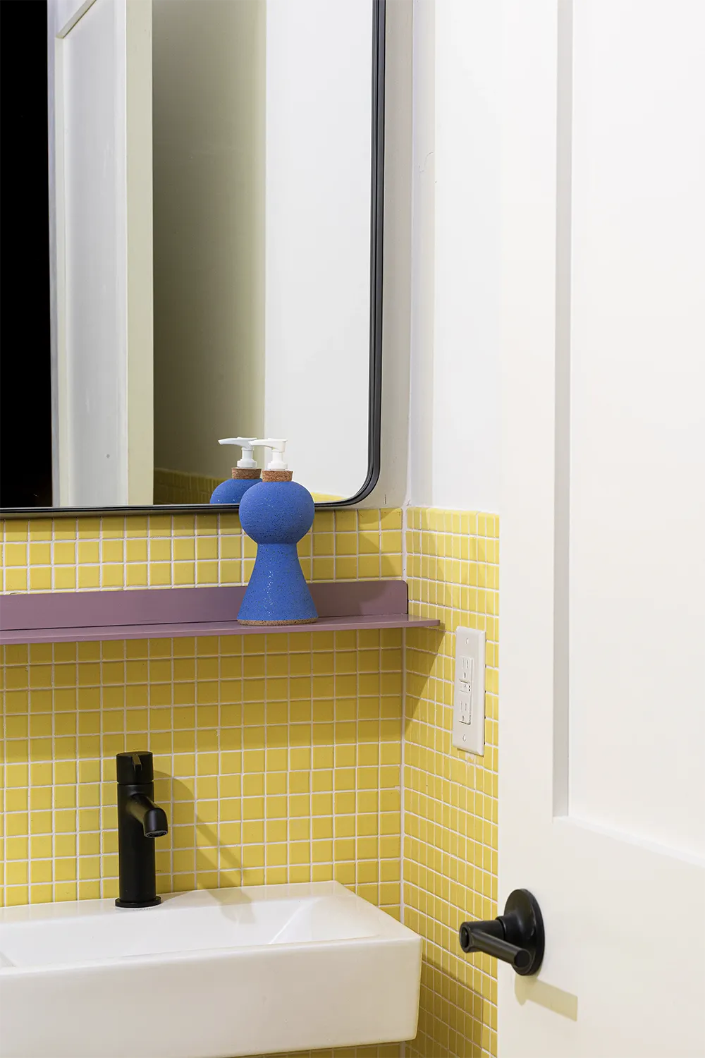 Bathroom corner with a blue hand soap dispenser on a purple shelf, a white sink in front of a yellow tiled wall