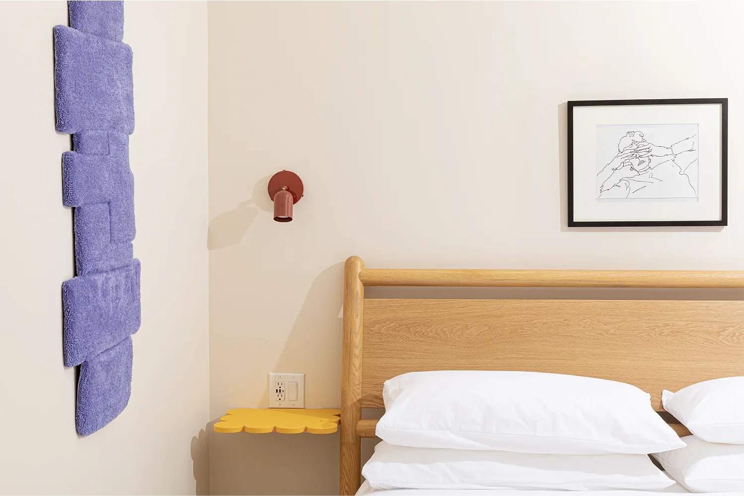 Beige bedroom with a wooden double bed with white bedding, a yellow floating bedside shelf, and a purple doormat frame hanging on the wall.