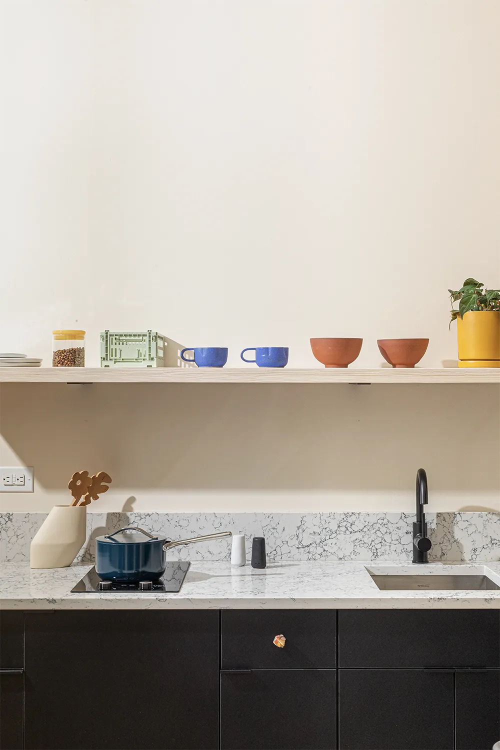 marble kitchen counter with sink and a pot, a beige shelf on top with blue mugs, orange bowls and a plant on a yellow flowerpot