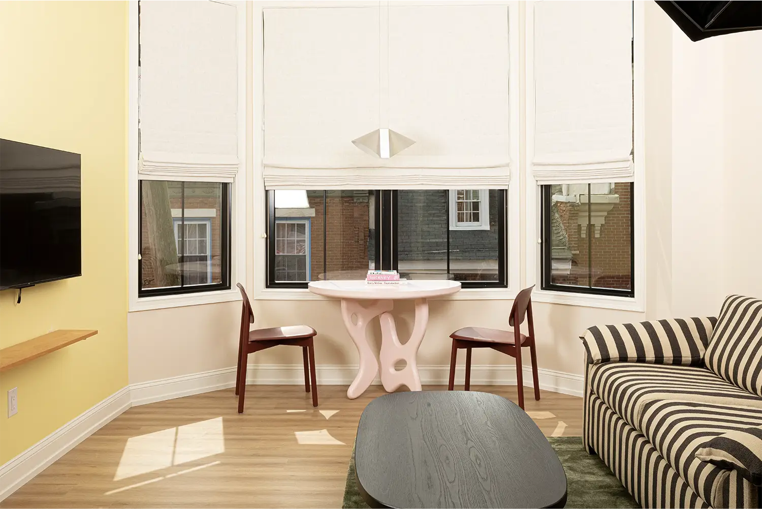 Beige and yellow living room whit an artistic pink table and two brow chairs next to a big set of three windows, a striped white and black sofa and a black coffee table