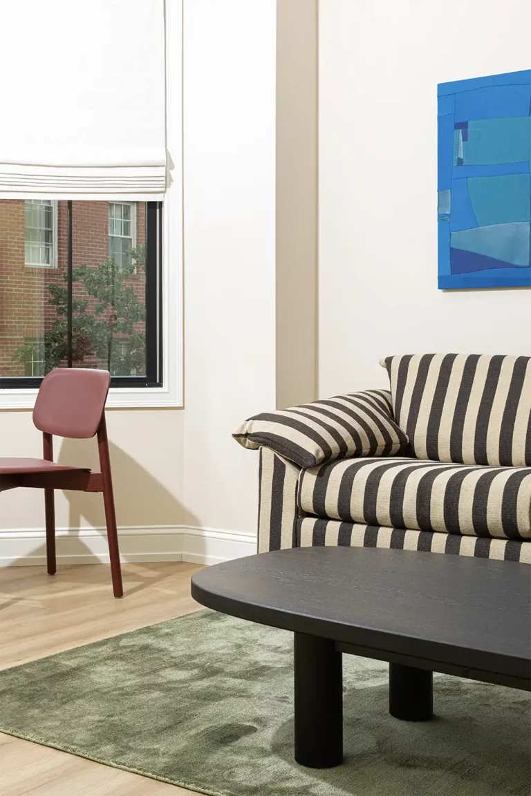 Corner of a beige room with a stripped black and white sofa, a blue painting on the wall behind, and a red wine chair next to a window