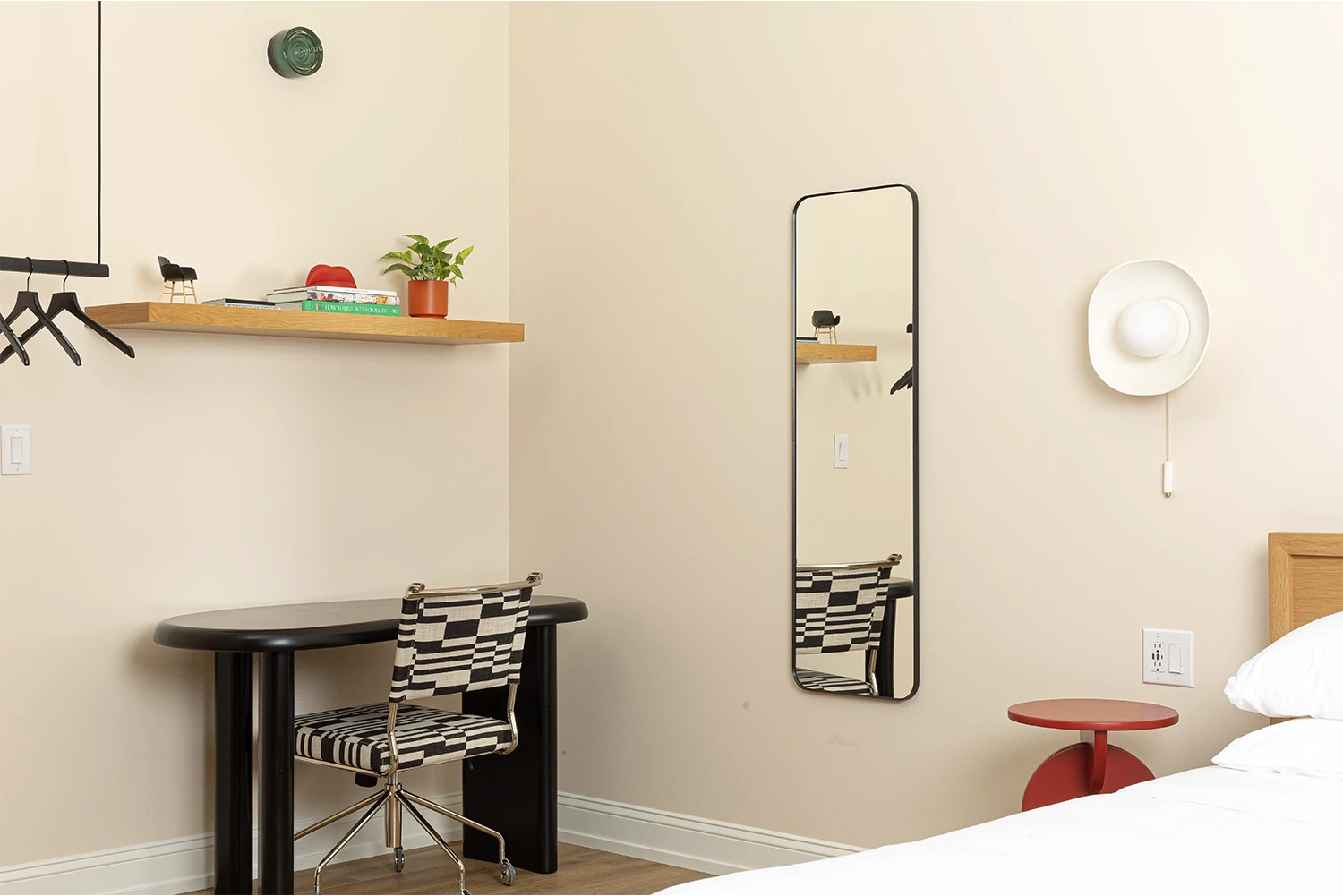 Beige bedroom view with a black desk and a white and black chair with a wood shelf whit books on it on top, a full-length mirror, a red nightstand and a white wall lamp above