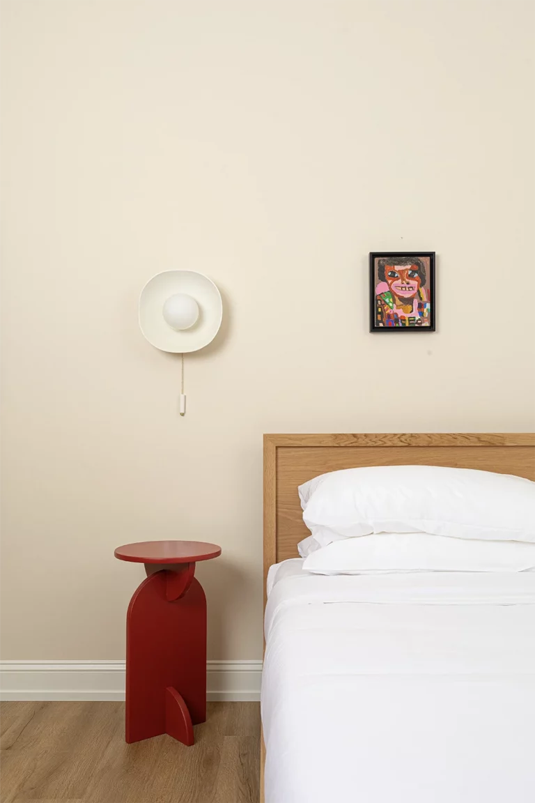 Wood bed whit white bedding, a small red nightstand, a round lamp above and a colorful picture