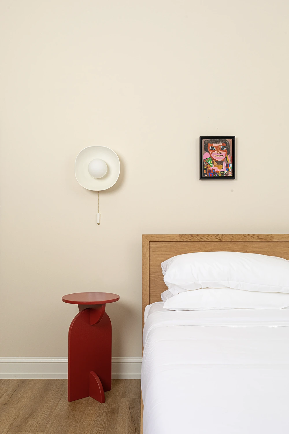 Wood bed whit white bedding, a small red nightstand, a round lamp above and a colorful picture