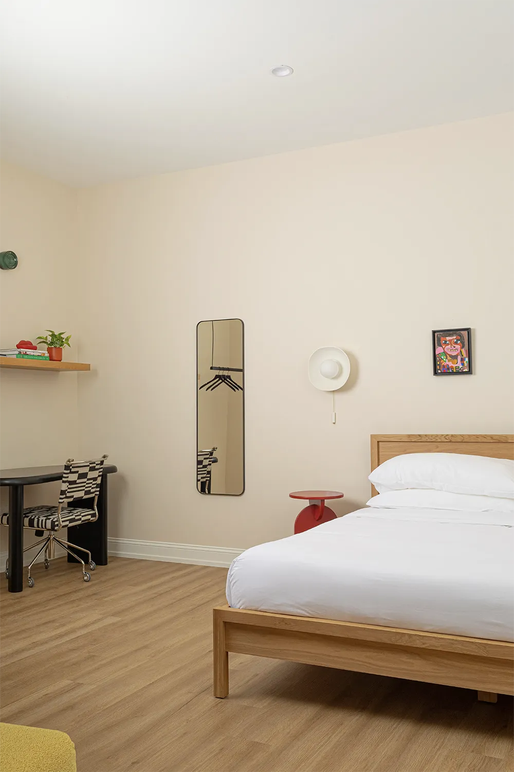 Beige bedroom view with a black desk and a white and black chair with a wood shelf whit books on it on top, a full-length mirror, a red nightstand and a white wall lamp above next to a double wood bed with white bedding