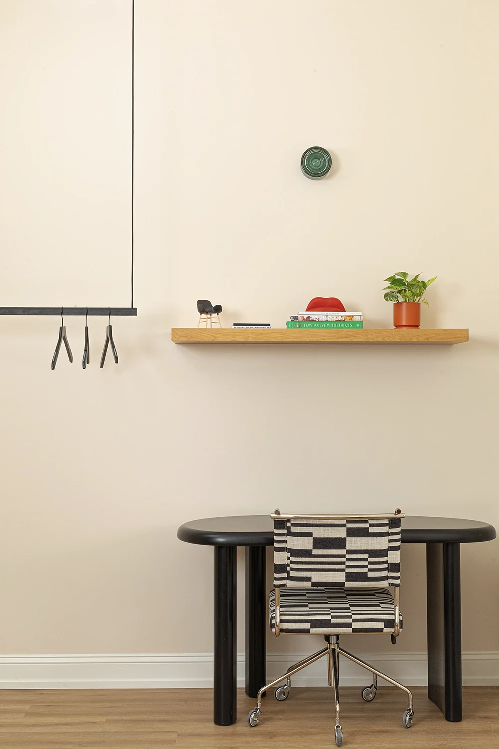 Black desk white a black and white chair, a wood shelf whit books and a plant on top, nex to it a hanging rack