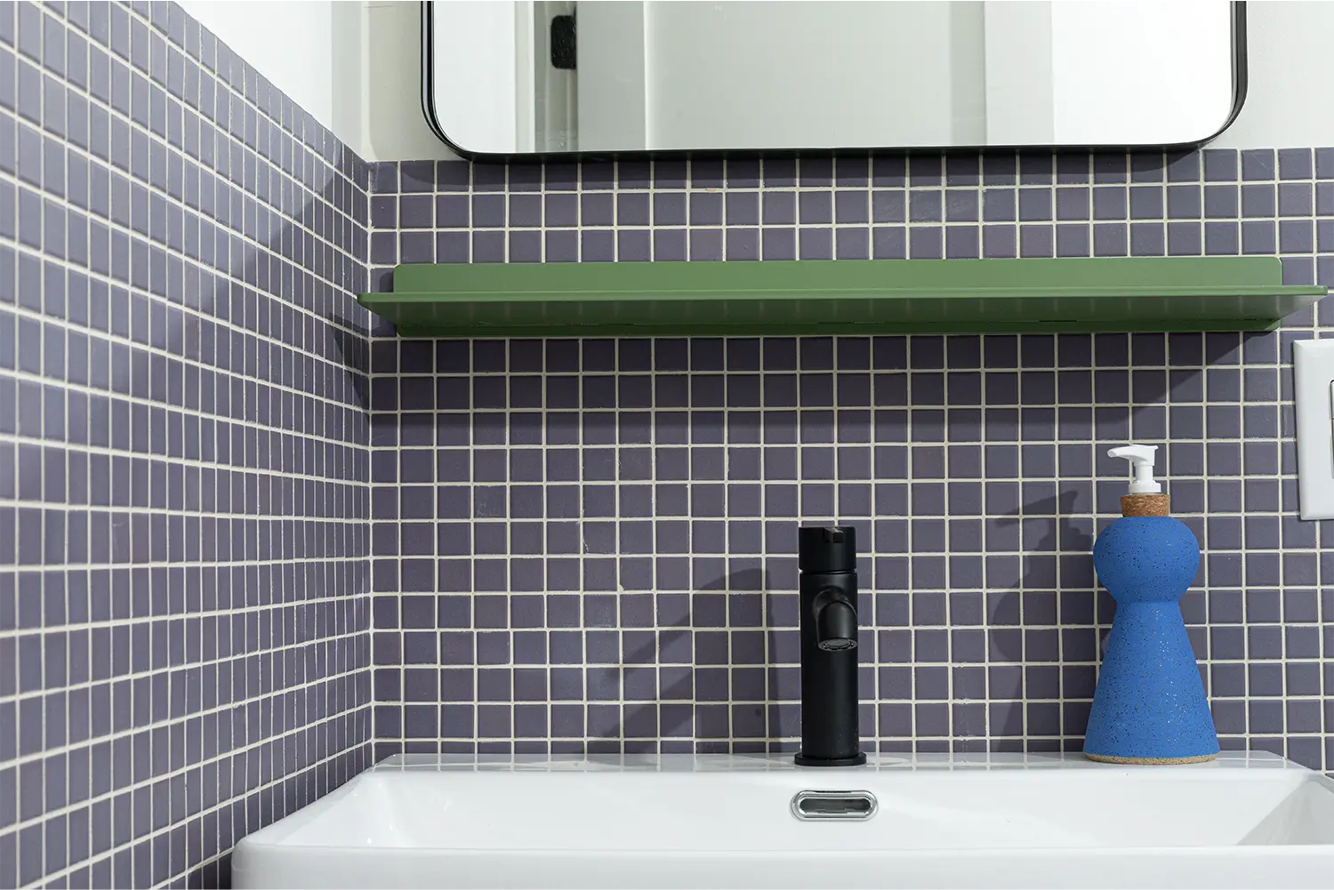 Bathroom sink with dark blue wall tiles, a green shelf and a blue hand soap dispenser