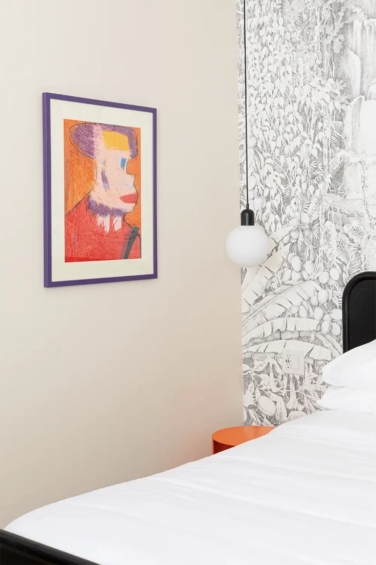 Beige room, with a bed with white sheets, a round hanging lamp, and a colorful painting