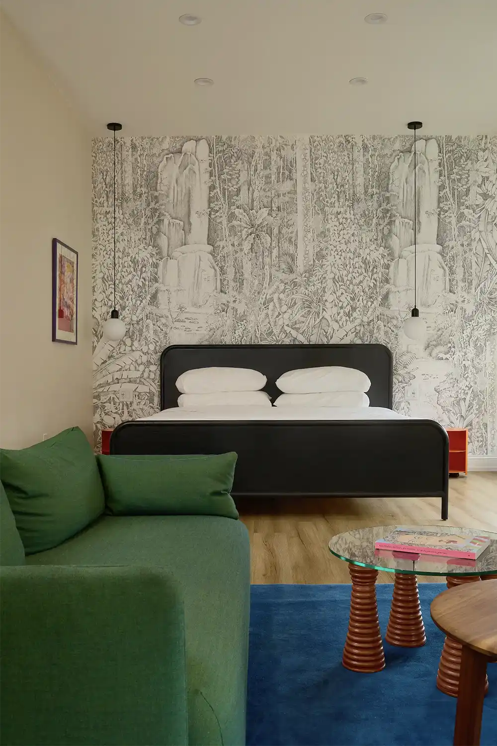 Beige walls bedroom with a black double bed on white sheets with two small hanging lamps on each side, a green sofa, wood and glass coffee table and blue carpet