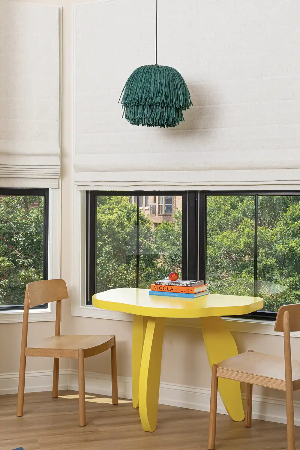 Artistic yellow table next to a large window, with two wooden chairs, books on top, and a hanging green lamp.