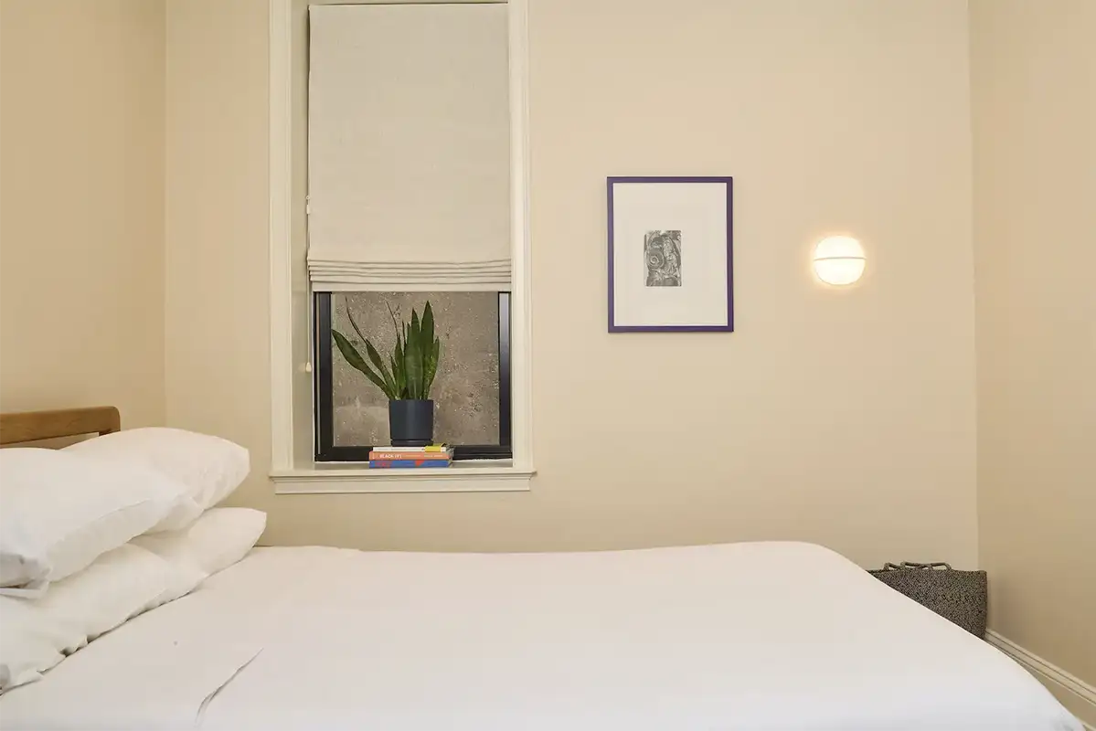 Beige bedroom whit a double bed next to a small window whit a plant on the frame