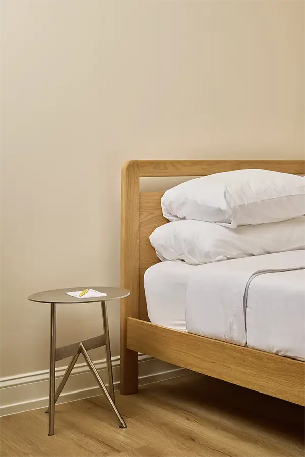 Minimalist bedroom with a wooden bed frame, white bedding, and a small round side table against a beige wall.