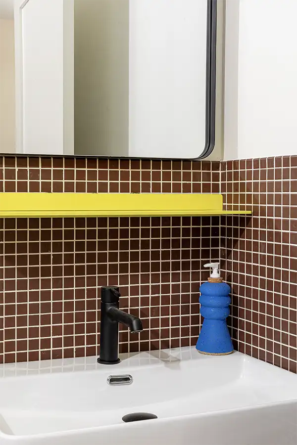 Bathroom corner with a blue hand soap dispenser on a white sink in front of a brown tiled wall with a yellow shelf above
