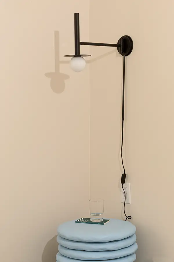 Corner of a beige room with an artistic light blue nightstand, a small boo, a glass of water on top and a black wall lamp above