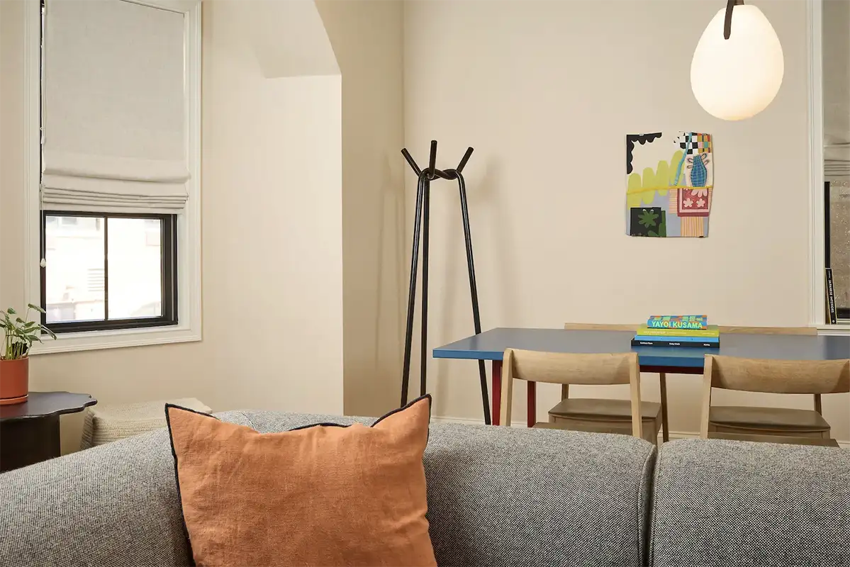 Blue wood table with 4 wood chairs around next to a window, books on top, a hanging lap and the back of a gray sofa