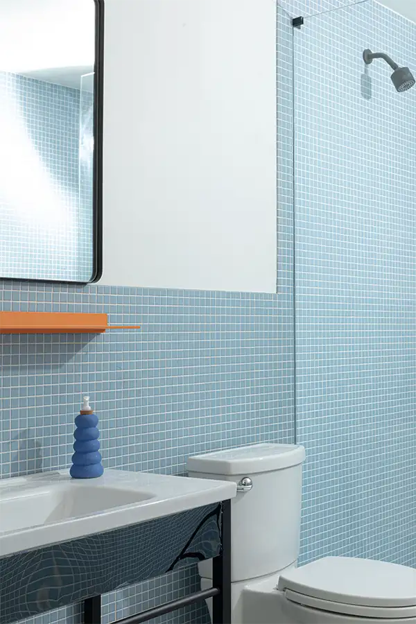 Bathroom with blue tiles, a large hanging mirror, a orange shelf underneath, a white sink with hand soap dispenser, a white toilet, and a glass-enclosed shower.