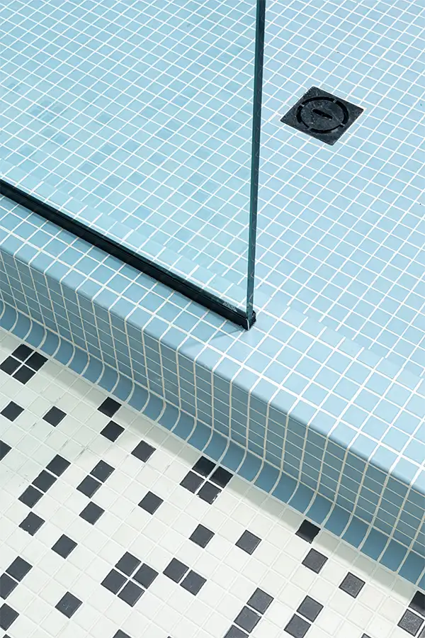 Modern bathroom design with pastel blue mosaic shower tiles and patterned black-and-white floor tiles, divided by a sleek glass panel.