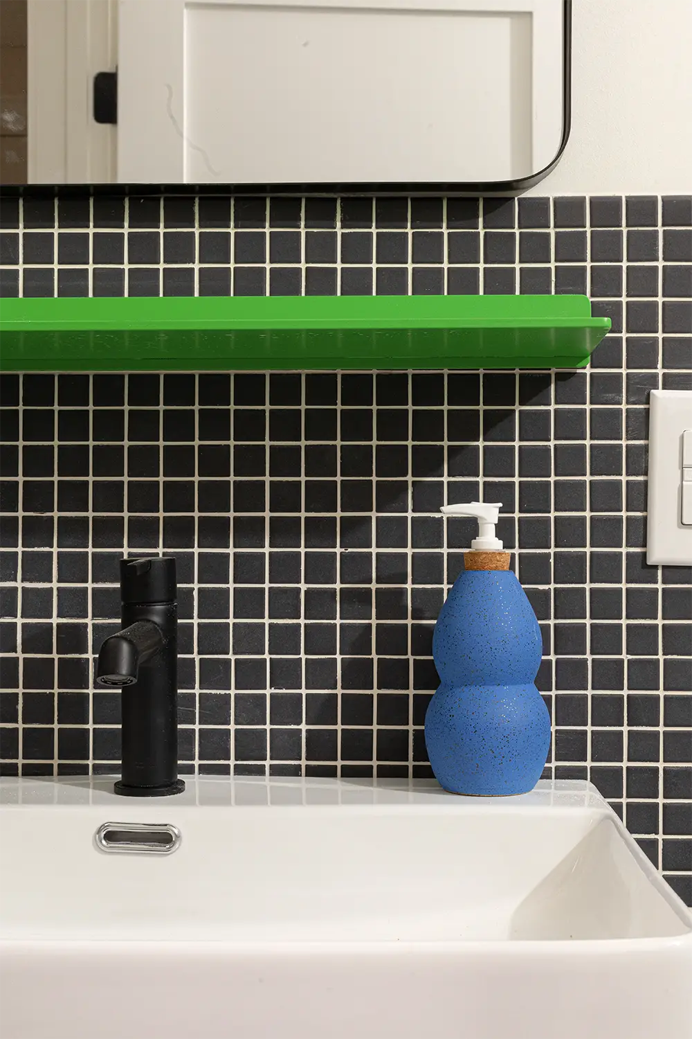 Bathroom corner with a blue hand soap dispenser on a white sink in front of a black tiled wall with a green shelf above