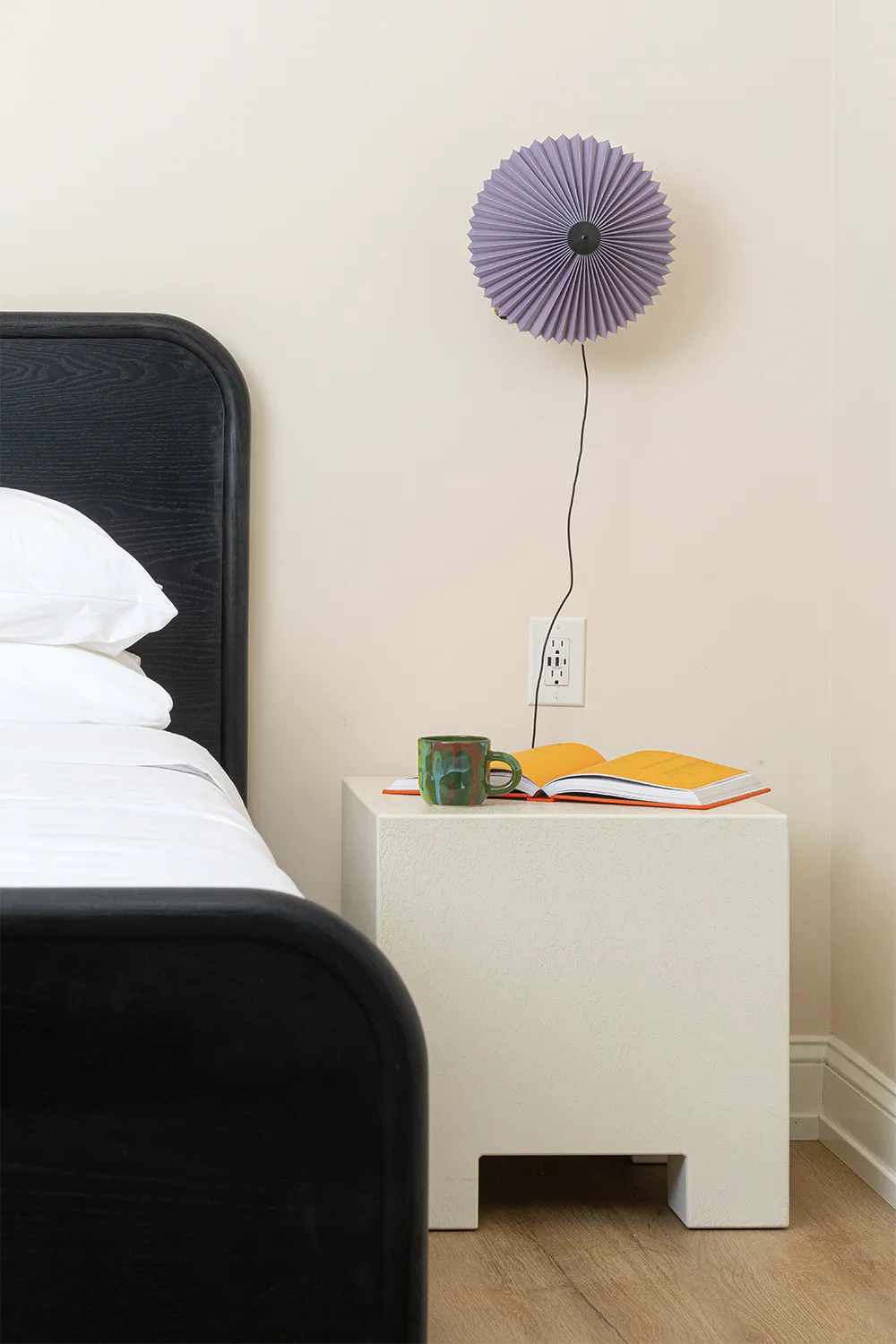 Corner of a beige room with a white nightstand with an open book and a green mug on top and an artistic purple wall lamp