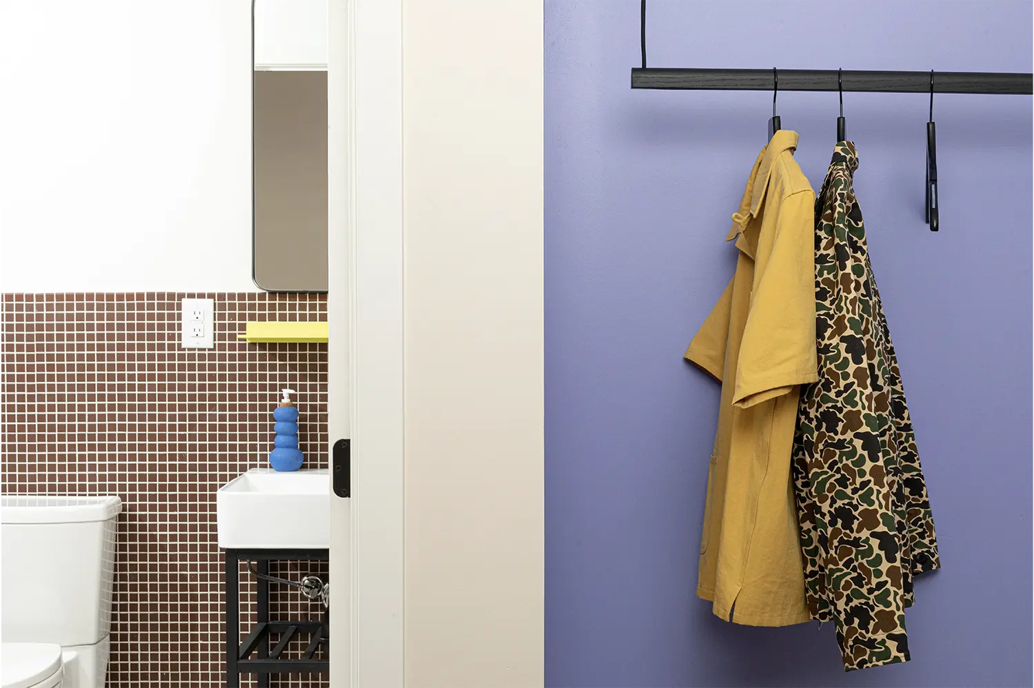Entrance of a bathroom next to a purple wall with a hanging rack with two colorful jackets on it