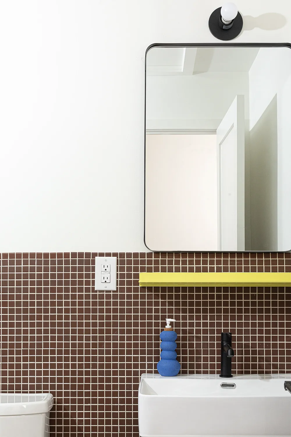 Bathroom with a blue hand soap dispenser on a white sink in front of a brown tiled wall and a mirror