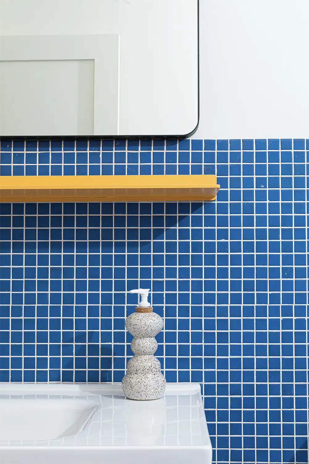 Bathroom with royal-style blue tiles, a large hanging mirror, and a yellow shelf below. White sink with hand soap dispenser.