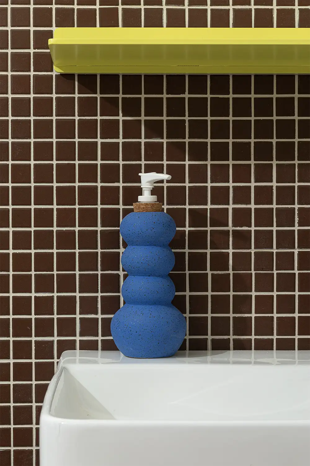 Blue hand soap dispenser on a white bathroom sink in front of a brown tiled wall