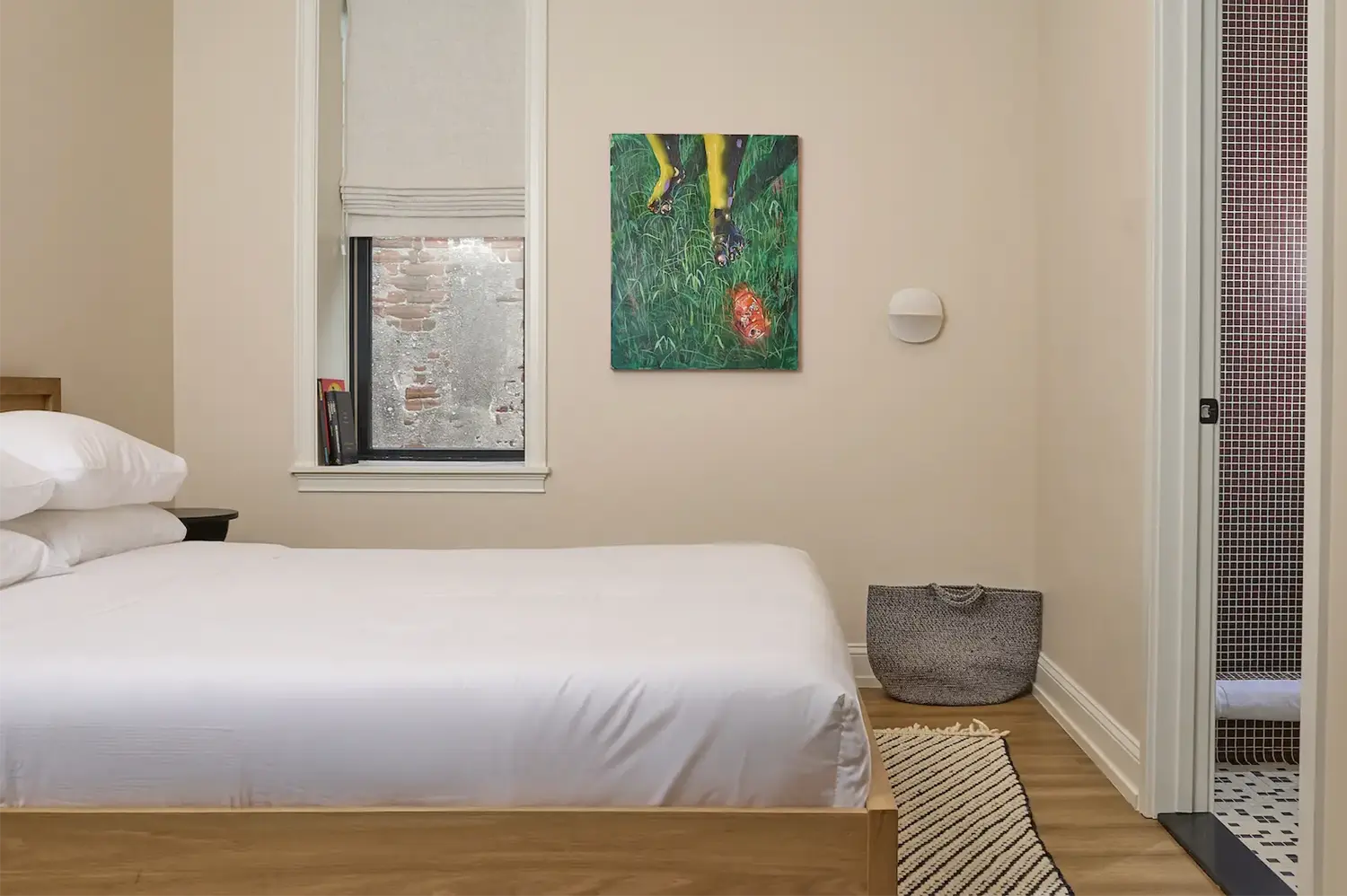 Beige room with a double wood foot bed with white sheets, a green painting hanging on the wall, a small window with off-white curtains and the door to the bathroom