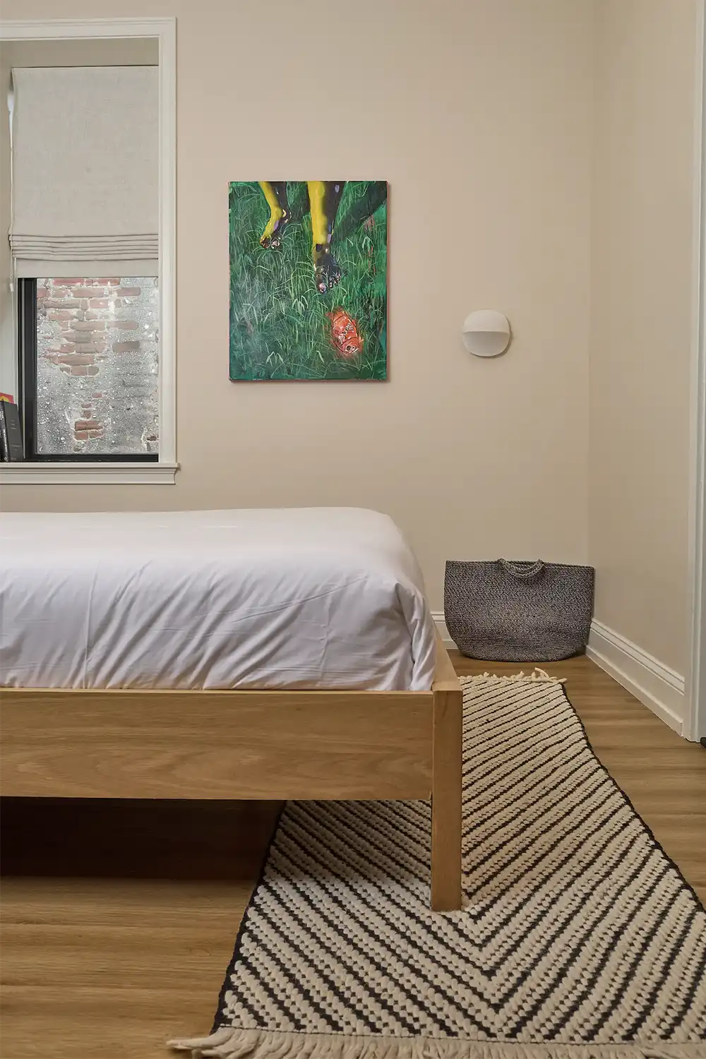 Beige room with a double wood foot bed with white sheets, a green painting hanging on the wall and a small window with off-white curtains