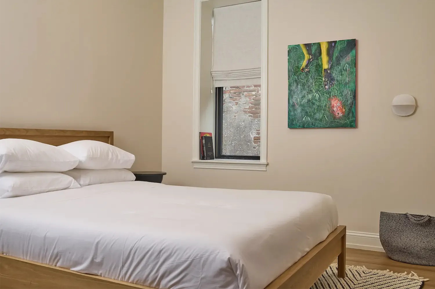 Beige room with a double wood bed with white sheets, a green painting hanging on the wall and a small window with off-white curtains