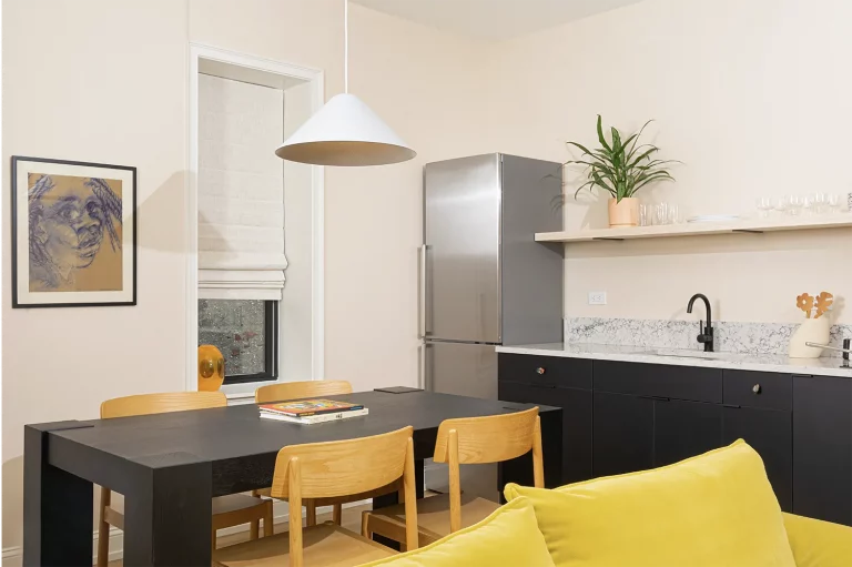 Corner of a kitchen with a silver refrigerator, a marble kitchen counter, a black table with wood chairs and a hanging white lamp