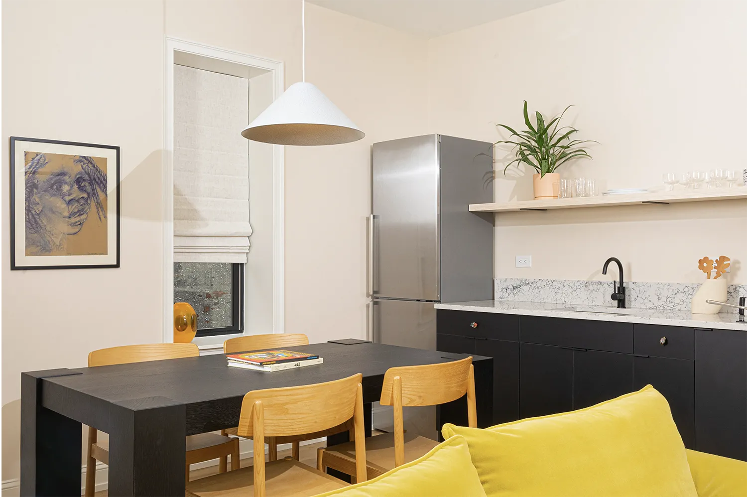 Corner of a kitchen with a silver refrigerator, a marble kitchen counter, a black table with wood chairs and a hanging white lamp