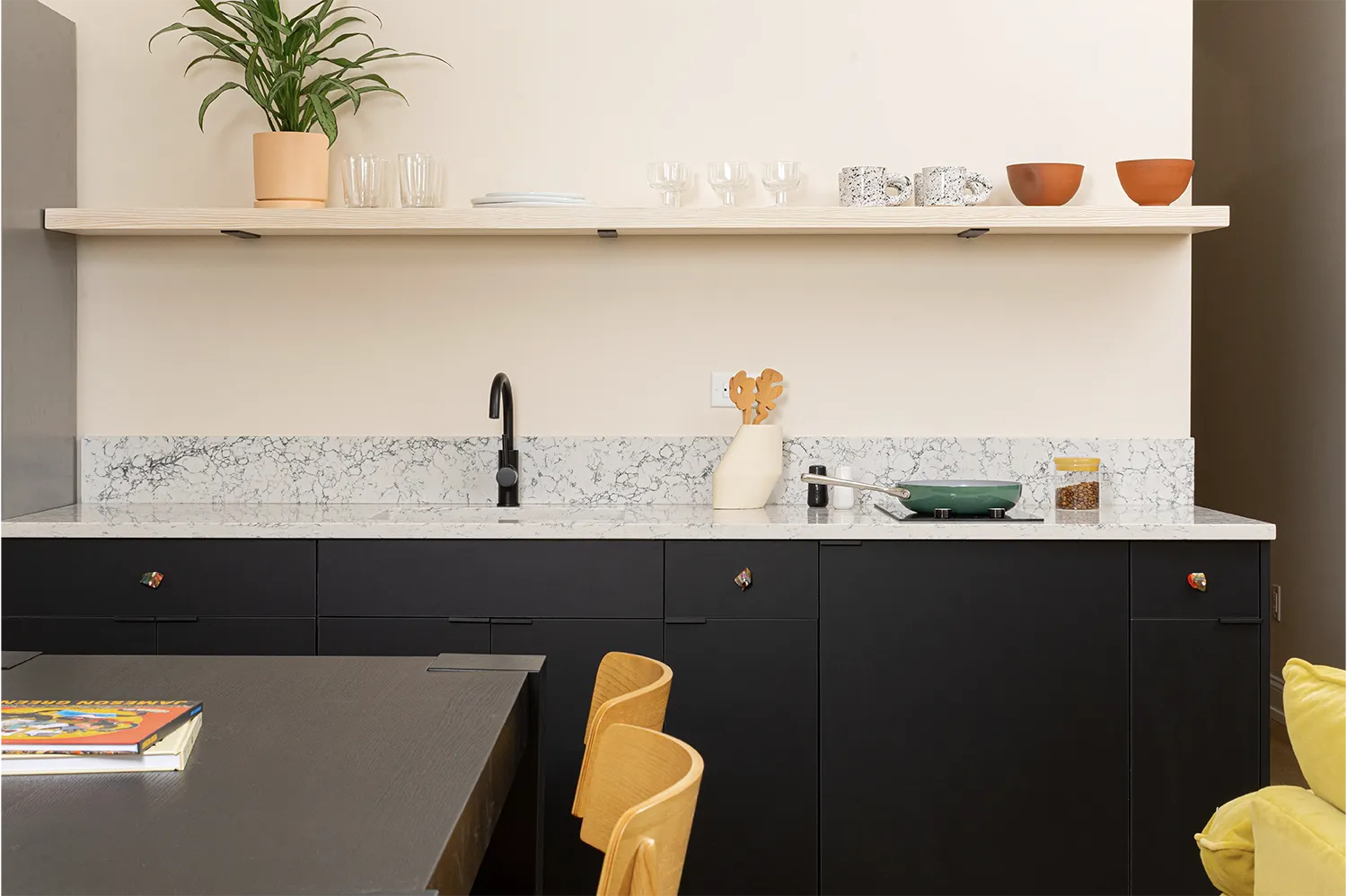 Marble kitchen counter with a pan on it and some utensils, a hanging beige shelf with cups, mugs and a green plant