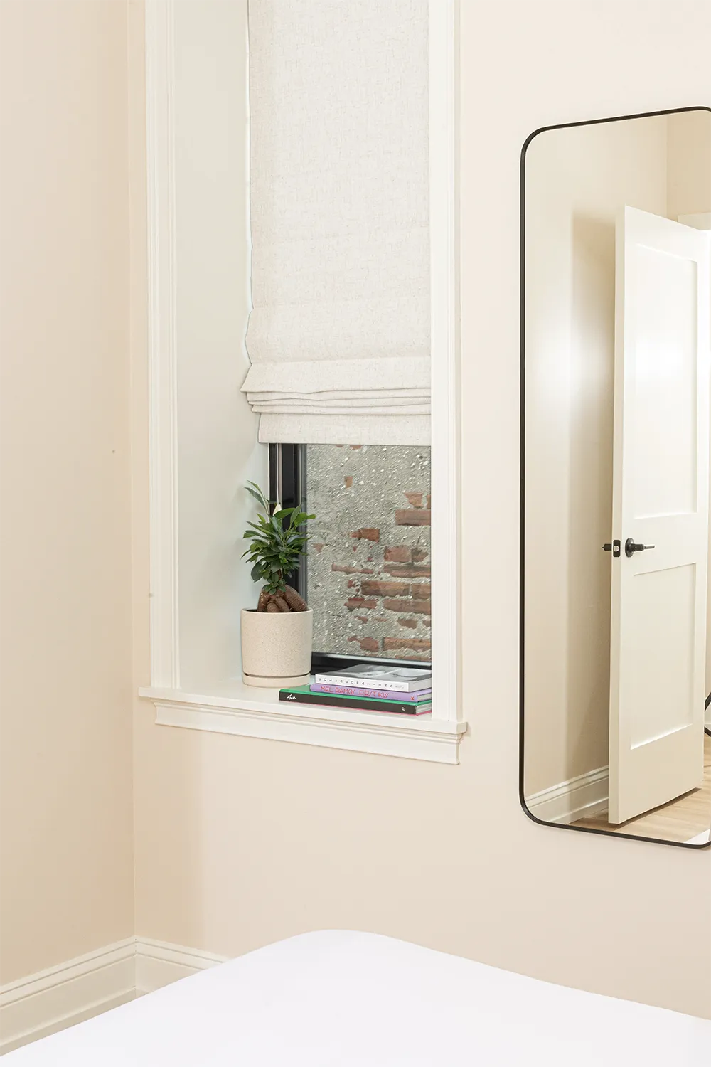 Corner of a beige room, a small window with off-white curtains and a full-length mirror