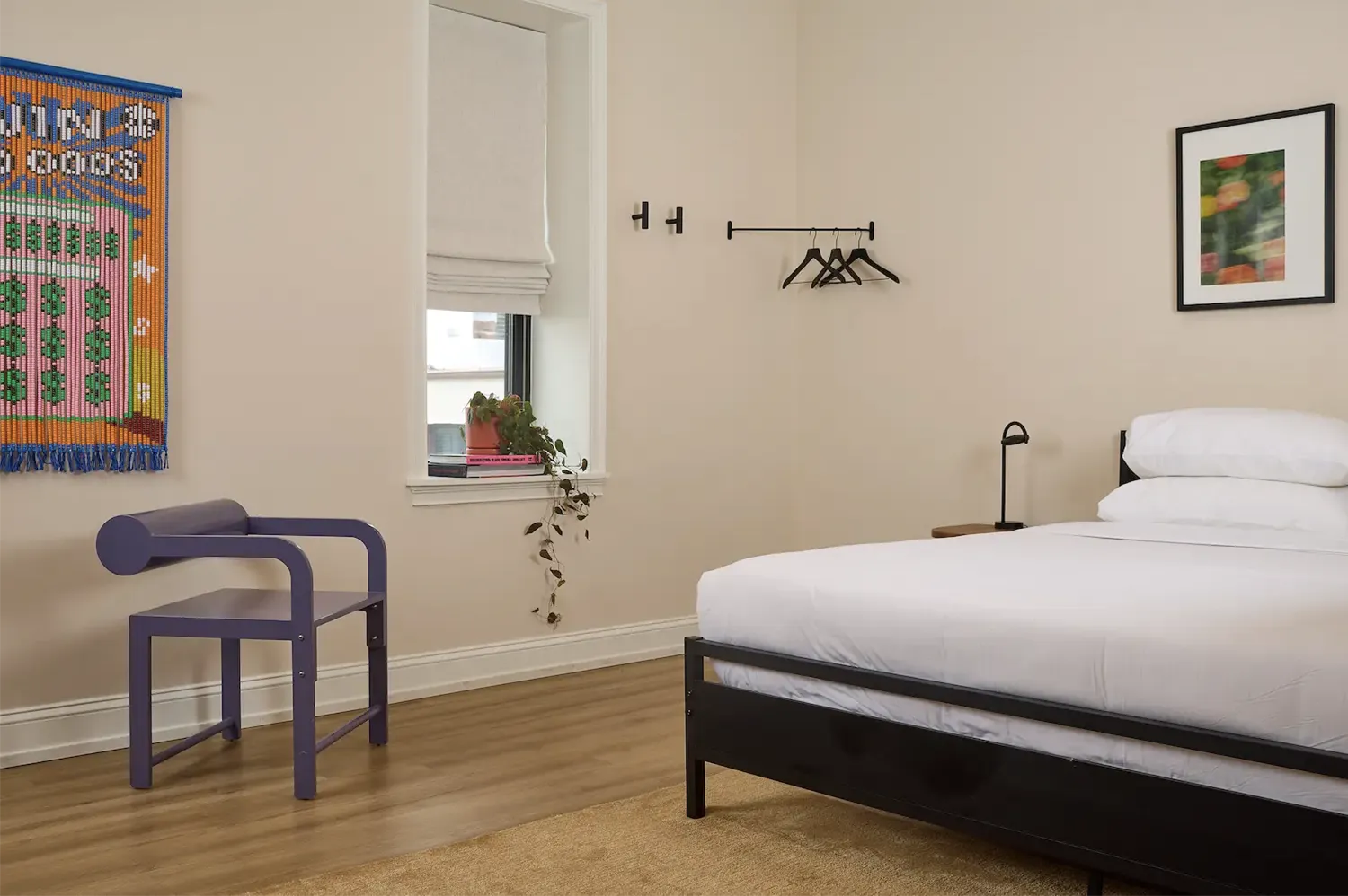 bedroom with a double bed with white bed sheets, a window frame whit a small plant, a picture on the wall and a purple chair