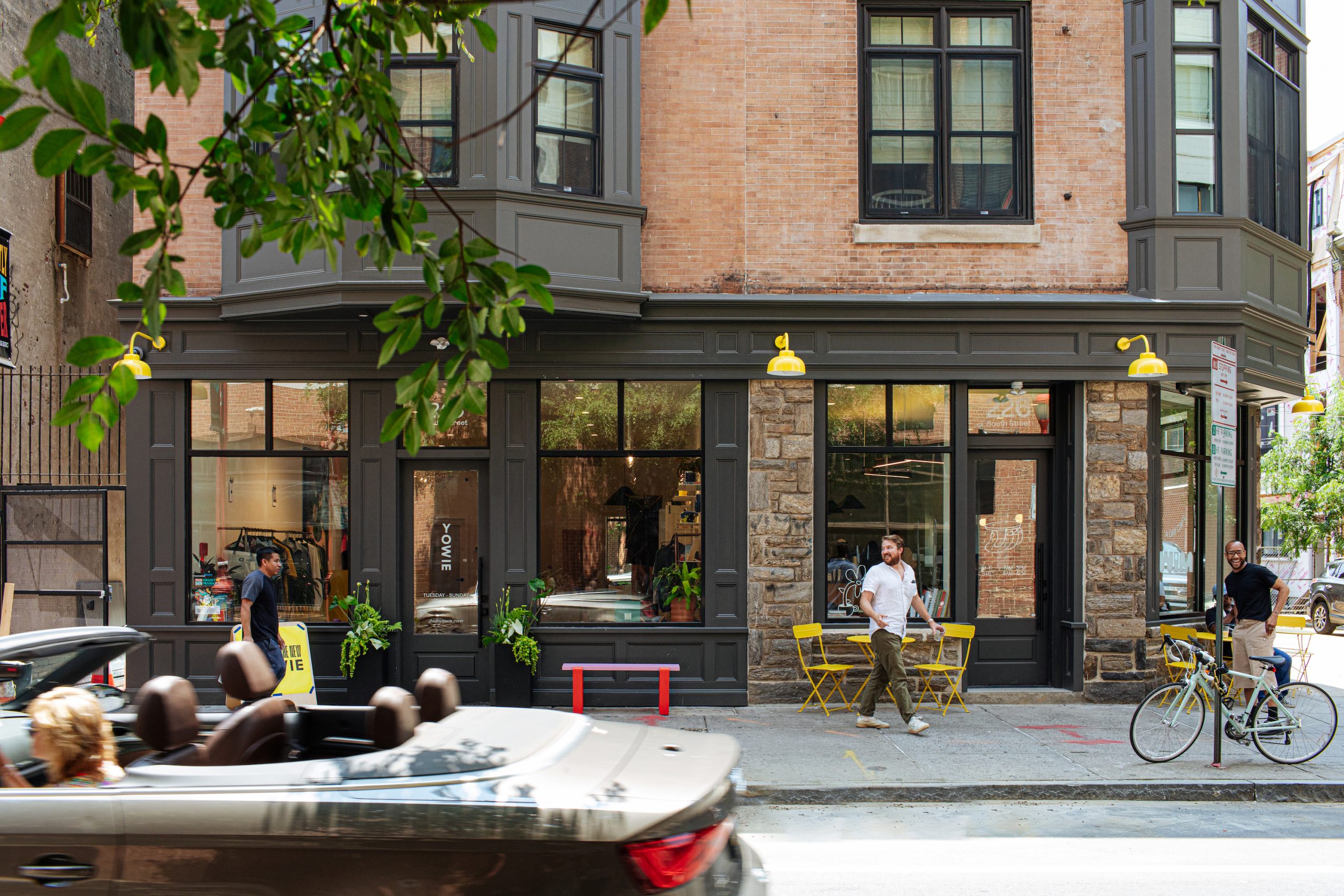 Street view of a corner entrance with large windows and gray trim on a brick building. Yellow chairs and tables are set outside, where one man is standing. Another man is walking past, while a bicyclist stands nearby. A car drives by in the foreground, and green tree branches frame the top left of the image.