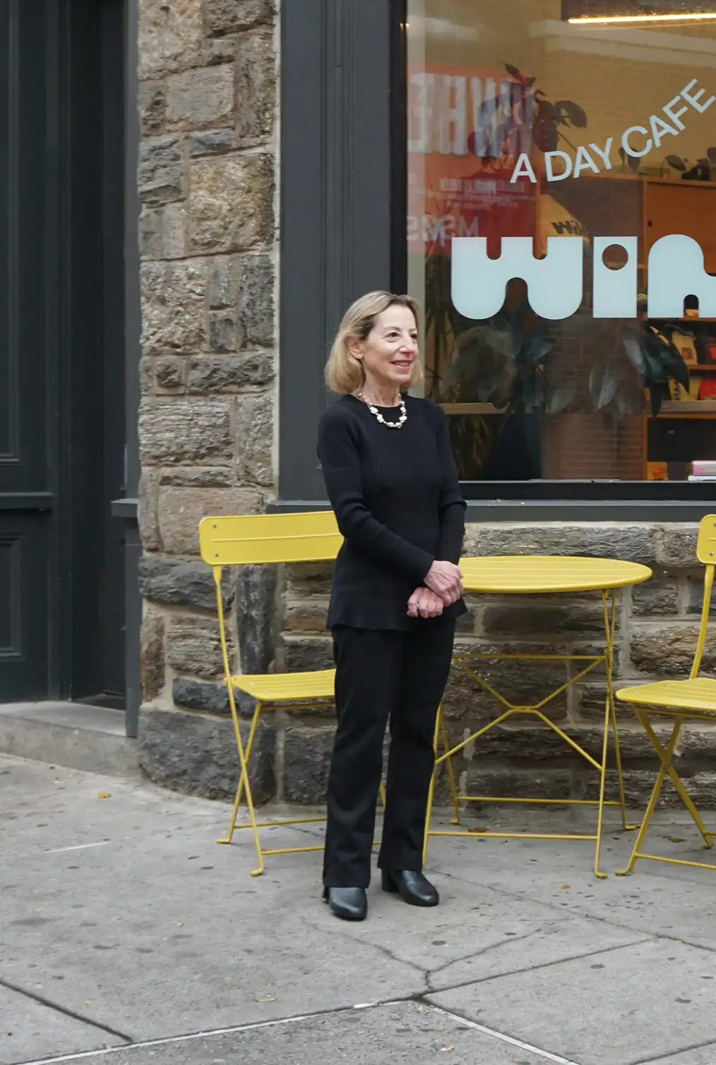 Adult woman dressed in black standing in front of a building with yellow chairs