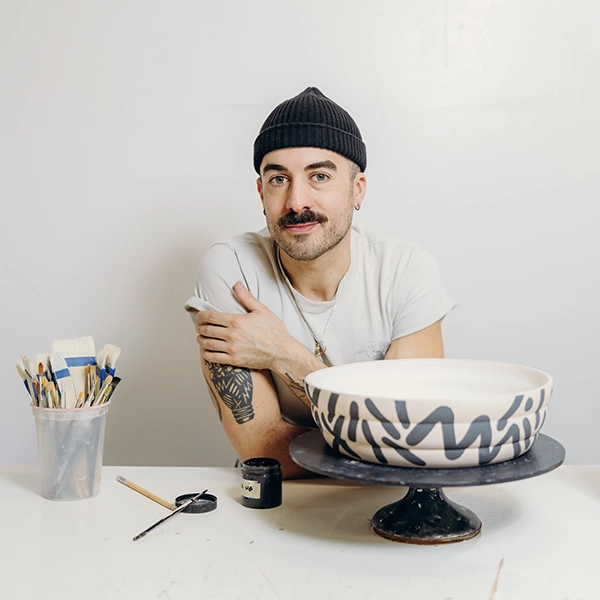 Portrait of a with man with green eyes and mustache wearing a white shirt and a beanie hat with a painted clay bowl, black paint and some brushes