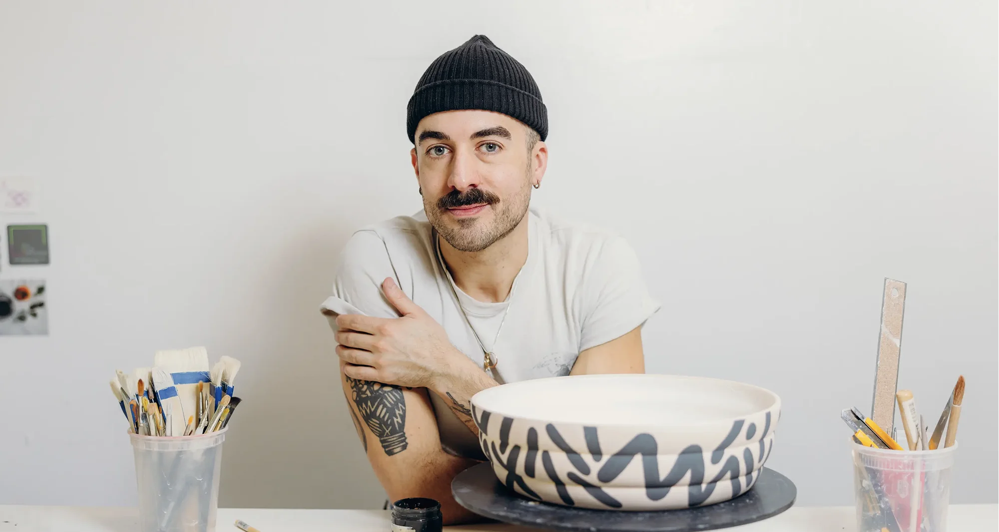 Portrait of a with man with green eyes and mustache wearing a white shirt and a beanie hat with a painted clay bowl, black paint and some brushes