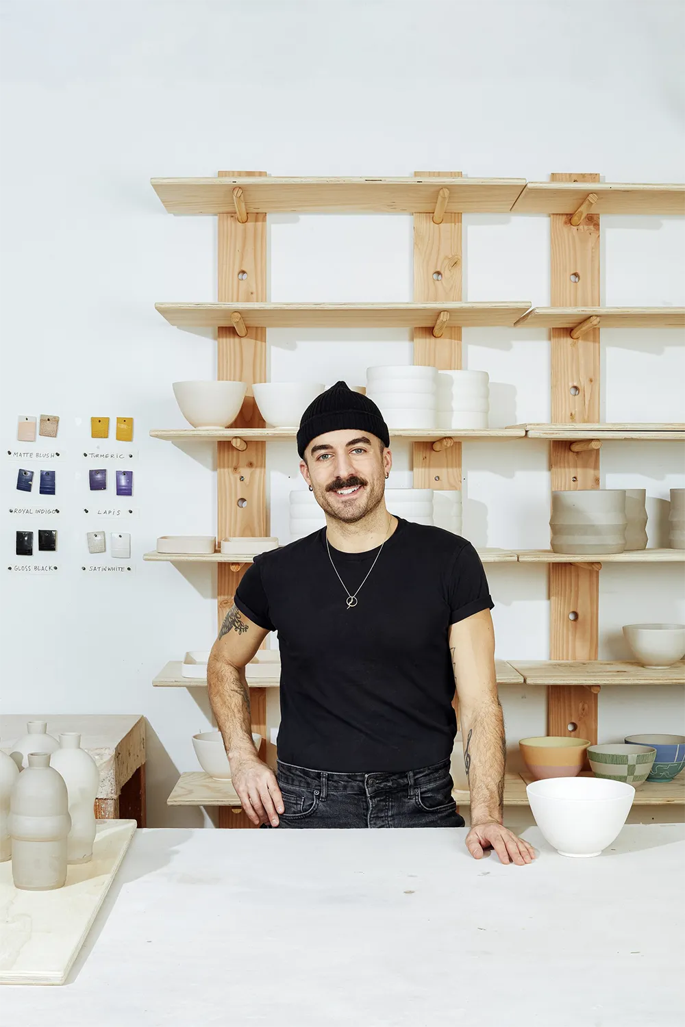 Portrait of a man with a mustache smiling, wearing black t-shirt, jeans and a beanie in front of a a stairwell full of ceramics